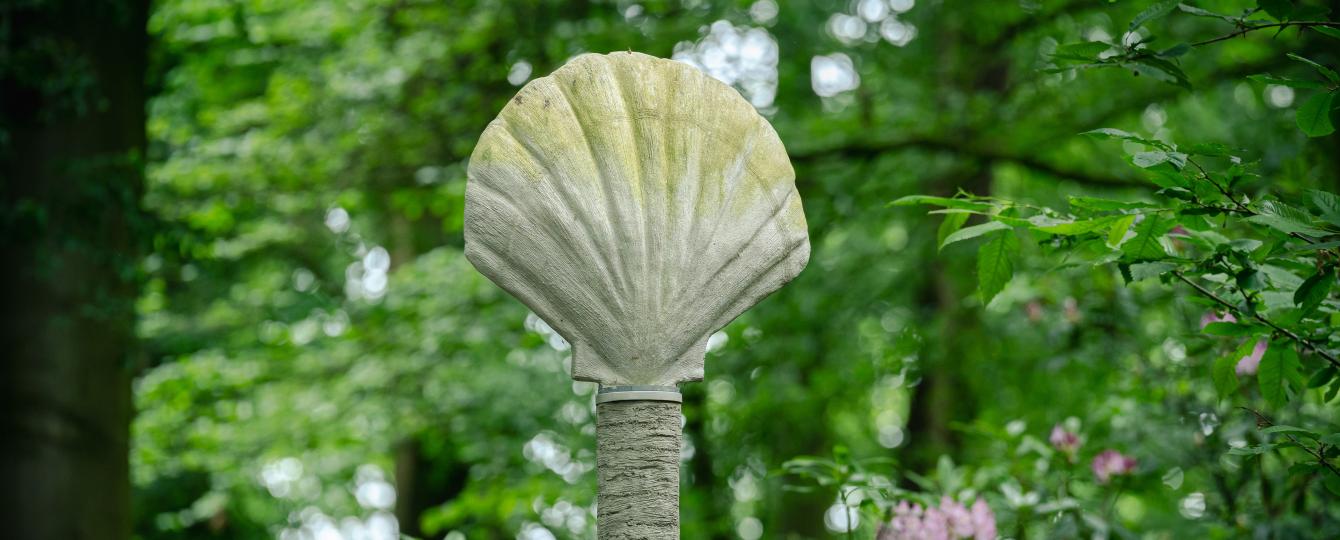 Schelpvormige sculptuur in rocaille op een hoge paal omgeven door een groene boomkruin