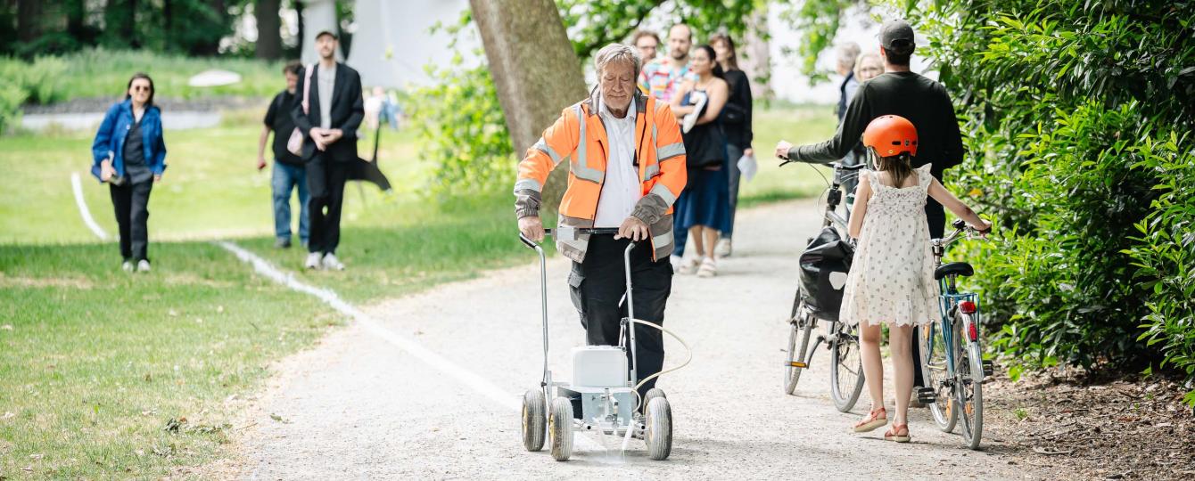 Erfgoedbewaker met fluohesje voert performance Le Tag van Joëlle Tuerlinckx uit en trekt een witte krijtlijn door het kunstpark terwijl bezoekers hem passeren.