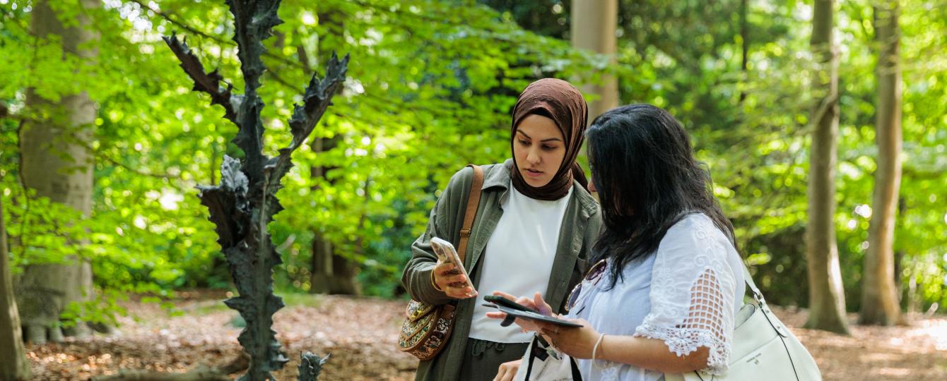 Jonge vrouw met hoofddoek en andere jonge vrouw kijken samen op smartphone en staan naast beeldhouwwerk in het kunstpark van het Middelheimmuseum.