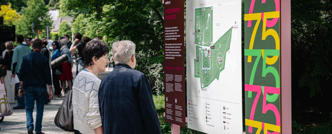 Koppel van middelbare leeftijd kijkend naar infobord aan hoofdingang kunstpark met op de achtergrond een aantal bezoekers