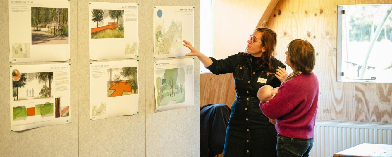 Sfeerbeeld van medewerker die uitleg geeft bij infopanelen met prints aan bezoeker met baby op de arm in Atelier van Lieshout.