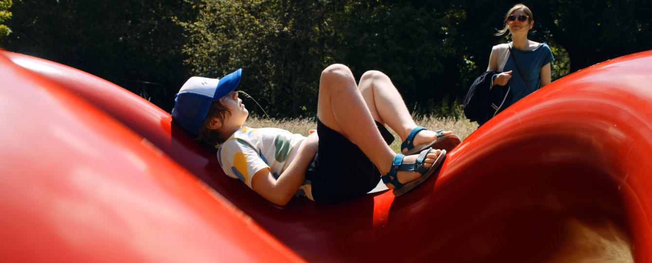 Jonge in zomerse outfit ligt op het rode kunstwerk Play Sculpture van Isamu Noguchi in het kunstpark van het Middelheimmuseum.