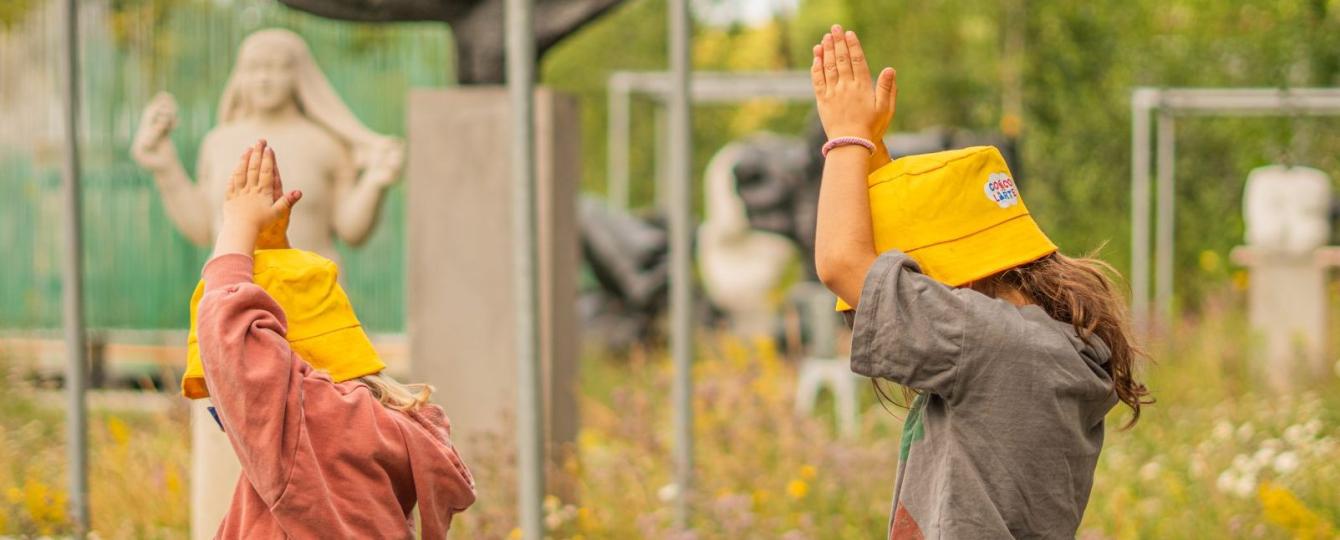 Twee jonge kinderen in zomeroutfit met gele zonnehoedjes wandelend met de armen boven het hoofd in het openluchtdepot van het Middelheimmuseum