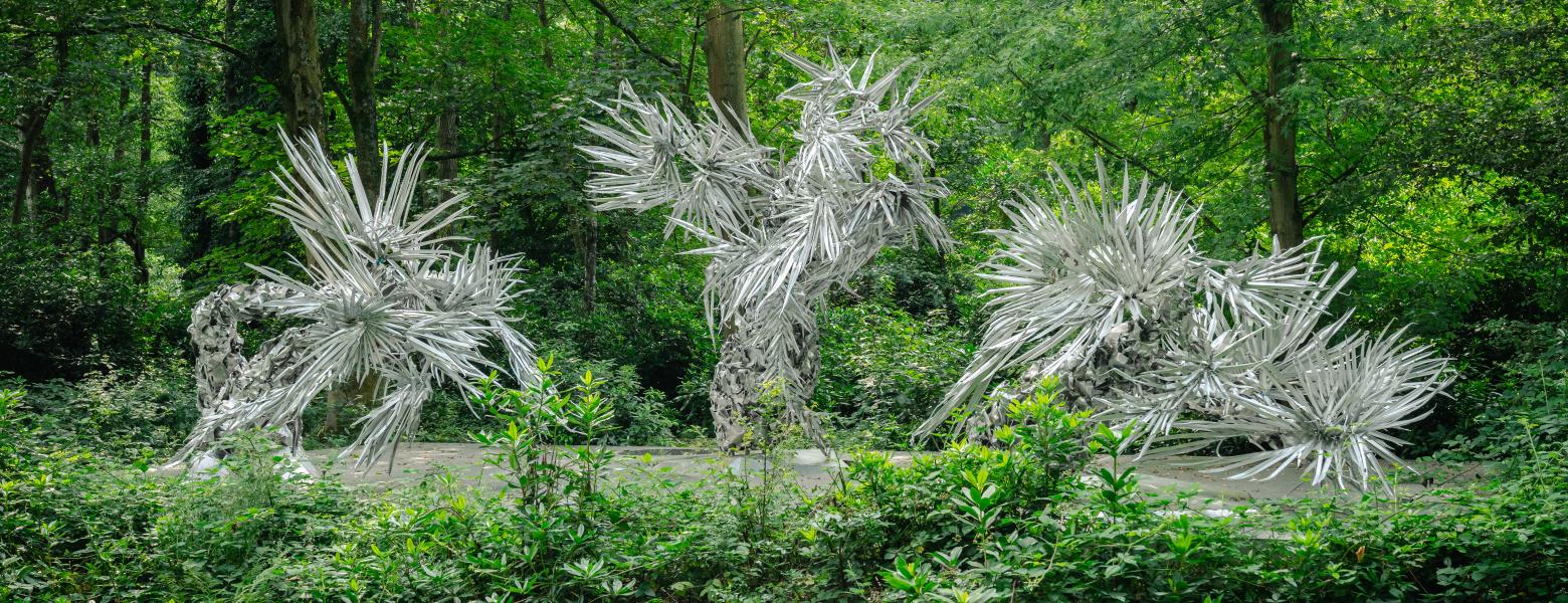 Frontaal zicht op de drie aluminium en roestvrij stalen palmboomachtige sculpturen omgeven door bomen en struiken