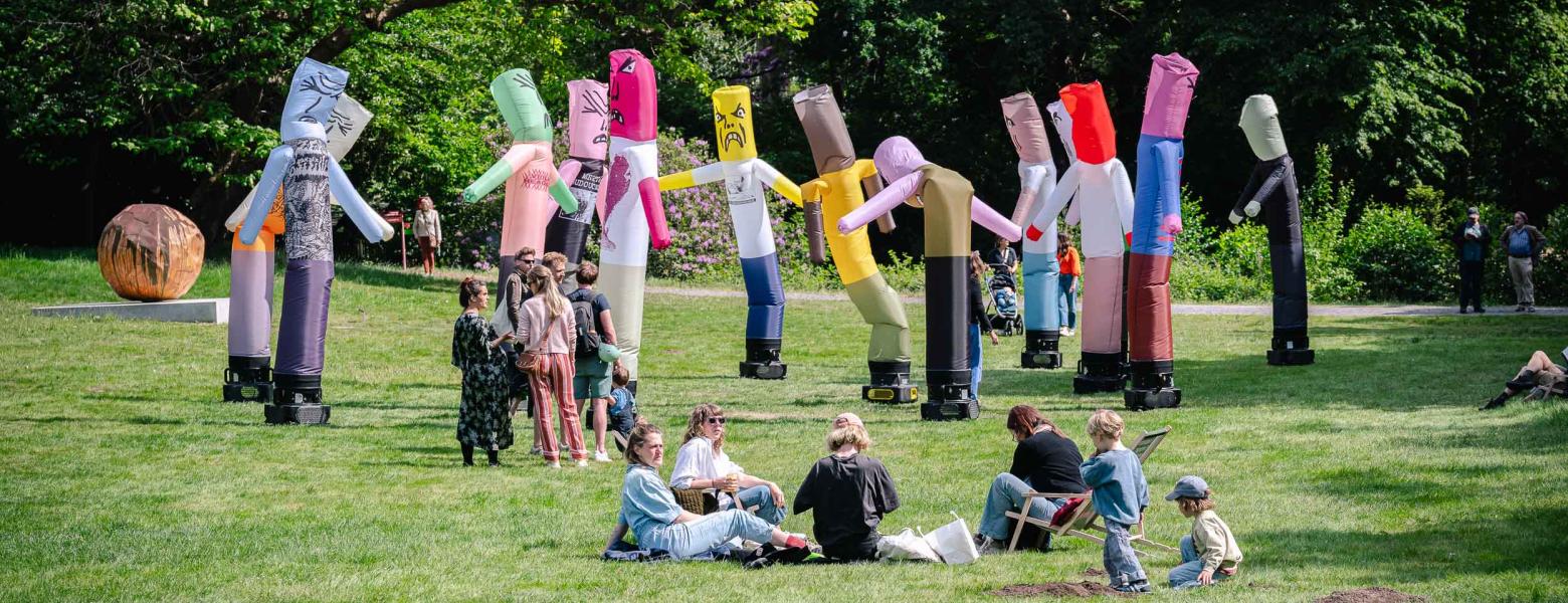Sfeerbeeld van groepje bezoekers zittend en groepje bezoekers staand op grasveld vlak voor de installatie met skytubes The Pogo Never Stops van Dennis Tyfus in het Middelheimmuseum.