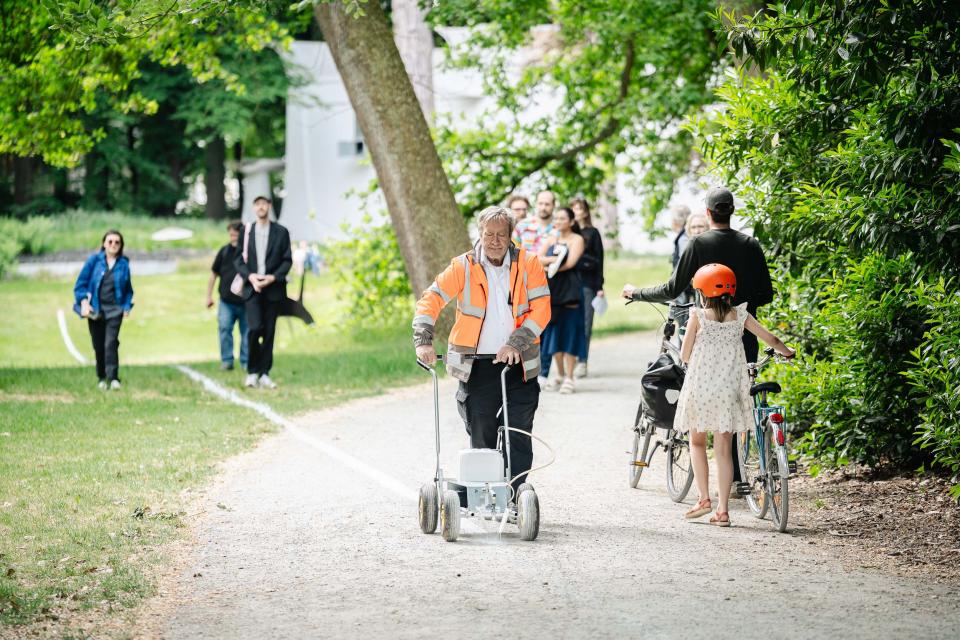 Erfgoedbewaker met fluohesje voert performance Le Tag van Joëlle Tuerlinckx uit en trekt een witte krijtlijn door het kunstpark terwijl bezoekers hem passeren.