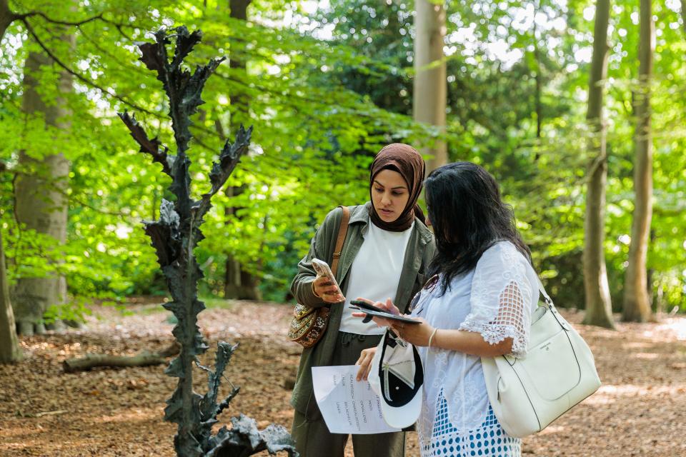 Jonge vrouw met hoofddoek en andere jonge vrouw kijken samen op smartphone en staan naast beeldhouwwerk in het kunstpark van het Middelheimmuseum.