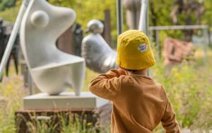 Kind in zomeroutfit en met geel zonnehoedje van Coccolarte wandelend langs het openluchtdepot in het kunstpark