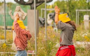 Twee jonge kinderen in zomeroutfit met gele zonnehoedjes wandelend met de armen boven het hoofd in het openluchtdepot van het Middelheimmuseum