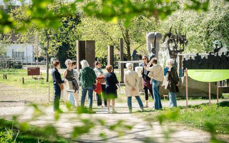 Zicht door het groene gebladerte op gids met groep aan het openluchtdepot van het Middelheimmuseum 