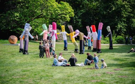 Sfeerbeeld van groepje bezoekers zittend en groepje bezoekers staand op grasveld vlak voor de installatie met skytubes The Pogo Never Stops van Dennis Tyfus in het Middelheimmuseum.