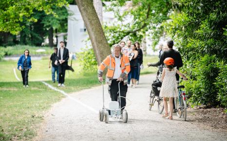 Erfgoedbewaker met fluohesje voert performance Le Tag van Joëlle Tuerlinckx uit en trekt een witte krijtlijn door het kunstpark terwijl bezoekers hem passeren.