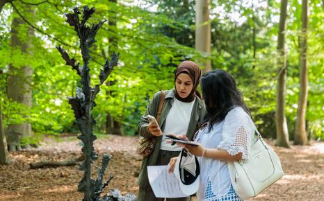 Jonge vrouw met hoofddoek en andere jonge vrouw kijken samen op smartphone en staan naast beeldhouwwerk in het kunstpark van het Middelheimmuseum.