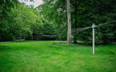 Zicht op grasveld met bomen en het kunstwerk Shouting is Breathing van Honoré d'O met paal en draden van en naar de bomen