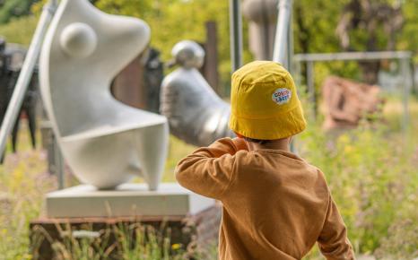 Kind in zomeroutfit en met geel zonnehoedje van Coccolarte wandelend langs het openluchtdepot in het kunstpark