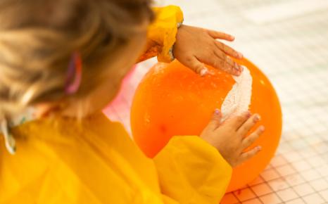 Sfeerbeeld van jong kind met gele schort en knutselend met oranje ballon en wit papier-maché