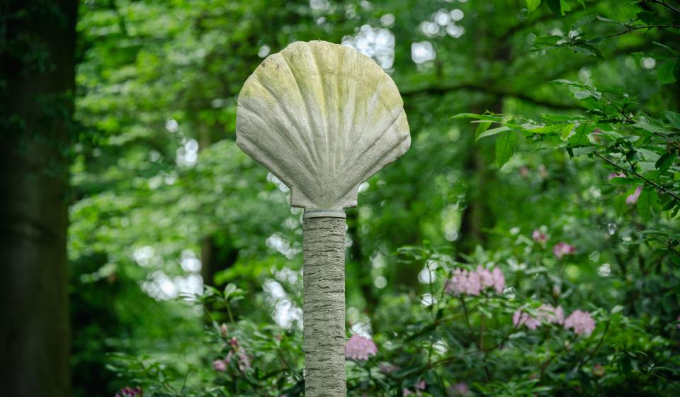 Schelpvormige sculptuur in rocaille op een hoge paal omgeven door een groene boomkruin