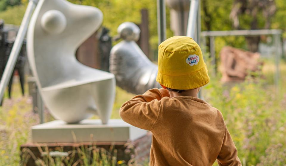 Kind in zomeroutfit en met geel zonnehoedje van Coccolarte wandelend langs het openluchtdepot in het kunstpark