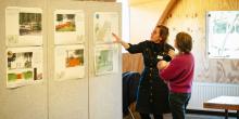 Sfeerbeeld van medewerker die uitleg geeft bij infopanelen met prints aan bezoeker met baby op de arm in Atelier van Lieshout.