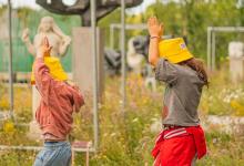 Twee jonge kinderen in zomeroutfit met gele zonnehoedjes wandelend met de armen boven het hoofd in het openluchtdepot van het Middelheimmuseum